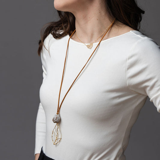 A woman wearing a white long-sleeve top and black pants, styled with a long gold pendant necklace and gold bracelets, photographed against a solid gray studio background.
