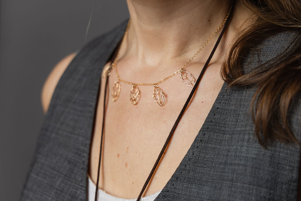 A close-up of a woman wearing a delicate rose gold leaf necklace layered over a dark brown cord necklace, set against a dark gray background.