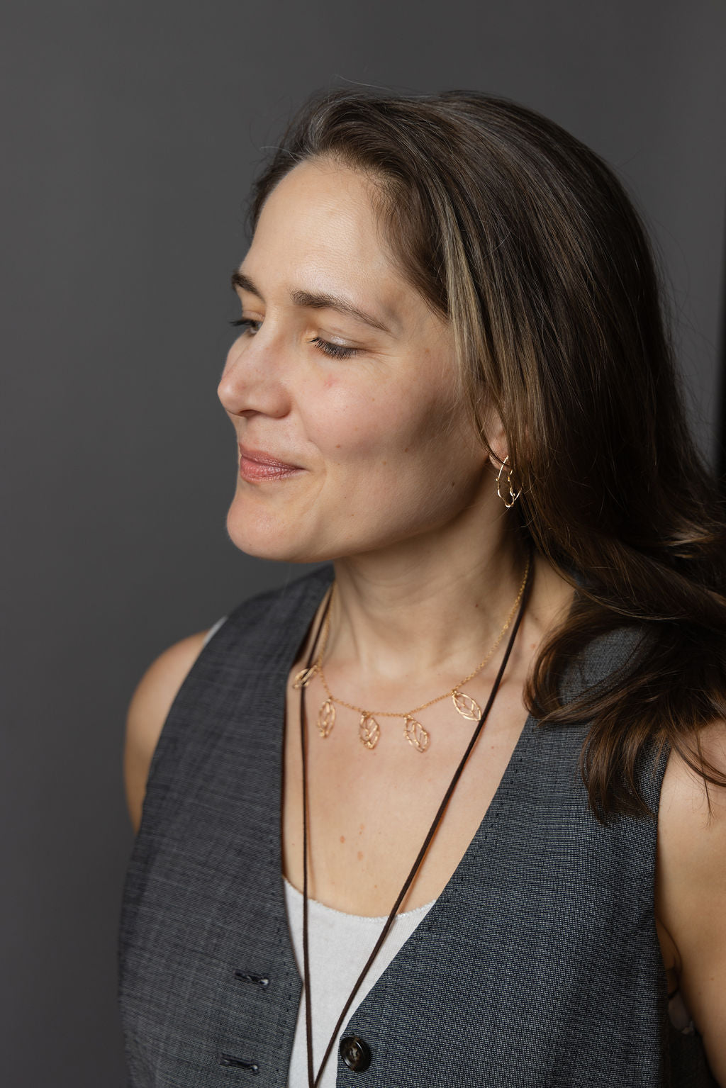 A woman in profile wearing a delicate rose gold necklace with small circular charms and matching rose gold hoop earrings, styled with a gray vest over a white top against a dark gray background.